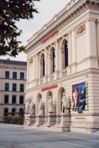 Side view of the Künstlerhaus with stone sculptures and colorful exhibition posters at street level.
