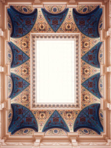 Ornate ceiling of the Künstlerhaus staircase hall, with geometric blue, gold, and terracotta patterns framing a central skylight.