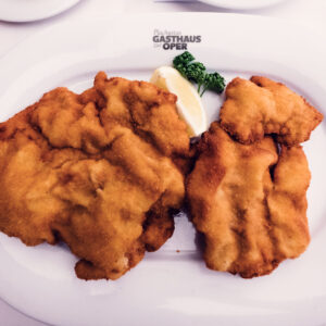 Wiener Schnitzel doré avec du citron et du persil, sur une assiette ovale portant le logo de Plachuttas Gasthaus zur Oper.