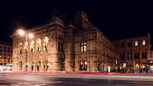 Wiener Staatsoper bei Nacht beleuchtet, goldenes Licht erzeugt dramatische Atmosphäre vor dem dunklen Himmel.
