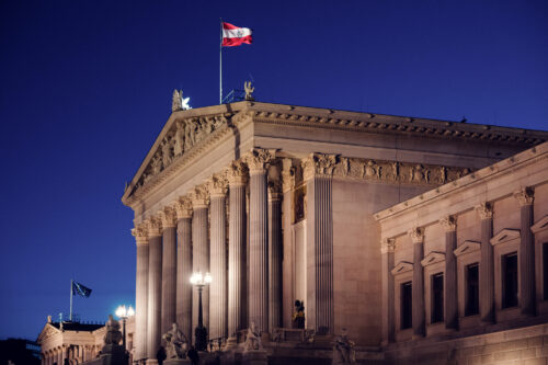El Parlamento austriaco de noche con columnas iluminadas y la bandera austriaca sobre un cielo azul oscuro.