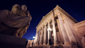 Figura de piedra sentada frente a las columnas iluminadas del Parlamento austriaco de noche.