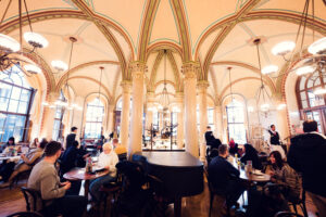 Vista grandangolare della sala principale del Café Central con ospiti ai tavoli rotondi sotto il soffitto a volta.