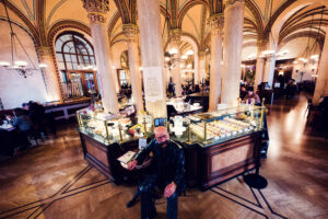 Interno del Café Central con alti soffitti a volta, colonne di marmo e il bancone delle paste con una statua di Peter Altenberg.