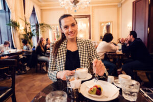 Adriana savourant un café et des pâtisseries dans le salon du Café Demel sous les lustres.