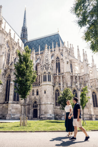 Adriana and Mario walking hand in hand along the stone pathway beside Votivkirche Vienna (Votive Church), with the neo-Gothic twin spires rising 99 meters against the summer sky.