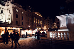 Visitors at the Christmas market am Hof at night, with the illuminated Marian Column visible in the foreground.