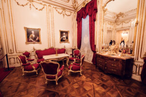 Barocker Konferenzraum im Schloss Schönbrunn mit roten Samtvorhängen und Möbeln aus der Epoche, kaiserliches Habsburger Palastinterieur mit prunkvollen hölzernen Stühlen um einen antiken Tisch, goldene Rokoko-Wanddekorationen und Porträtgemälde, Kristalllüster und Parkettboden.