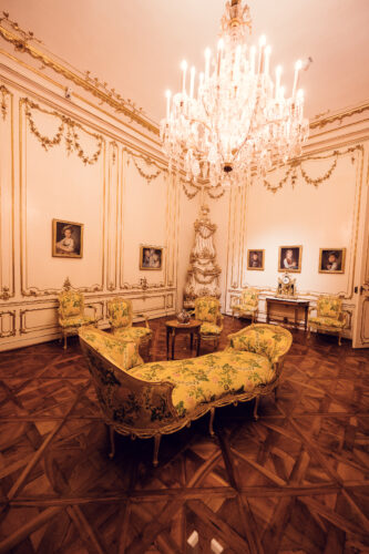 Luxuriöse Palaststube im Schloss Schönbrunn mit vergoldeten Rokoko-Wandpaneelen und historischen Möbeln, barockes österreichisches kaiserliches Interieur mit gelben Stühlen aus der Epoche und hölzernem Tisch, prunkvolle goldene Verzierungen und kristallene Beleuchtung.