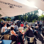 Belebte Terrasse des Palmenhaus mit Gästen unter Sonnenschirmen.