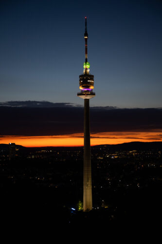 Vienna's Donauturm illuminated at night, its 252-meter structure glowing with LED lights against the dark sky.