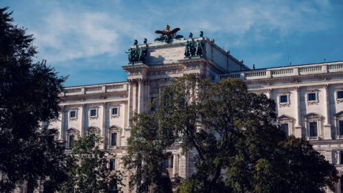 Neue Burg wing of the Hofburg seen from Burggarten, framed by trees.