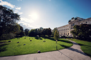 Amplio césped verde del Burggarten en un día soleado con la Neue Burg a lo lejos y visitantes sobre la hierba.