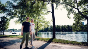 Adriana und Mario spazieren am Ufer der Alten Donau in Wien mit natürlicher Flusslandschaft und ruhigem Wasser im Hintergrund.