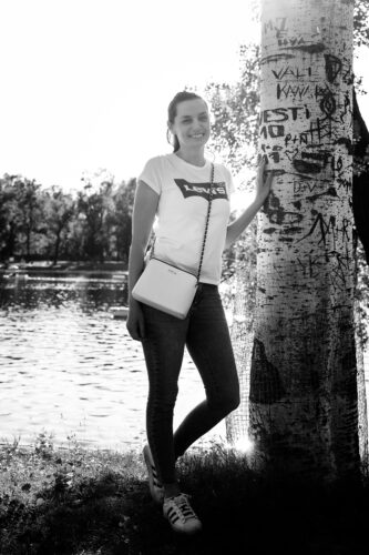 Portrait of Adriana at tree along Alte Donau riverbank in Vienna with tranquil water surface and shoreline vegetation behind.