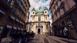 Blick von der Straße auf die Peterskirche in Wien mit der elliptischen Struktur, den beiden Glockentürmen und dem reich verzierten Eingangsportal mit Steinskulpturen.