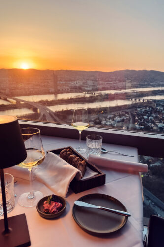 Sunset view from Donauturm's revolving restaurant at 170 meters height, overlooking Vienna's skyline with the meandering Danube River, Neue Donau, and the illuminated city lights below.