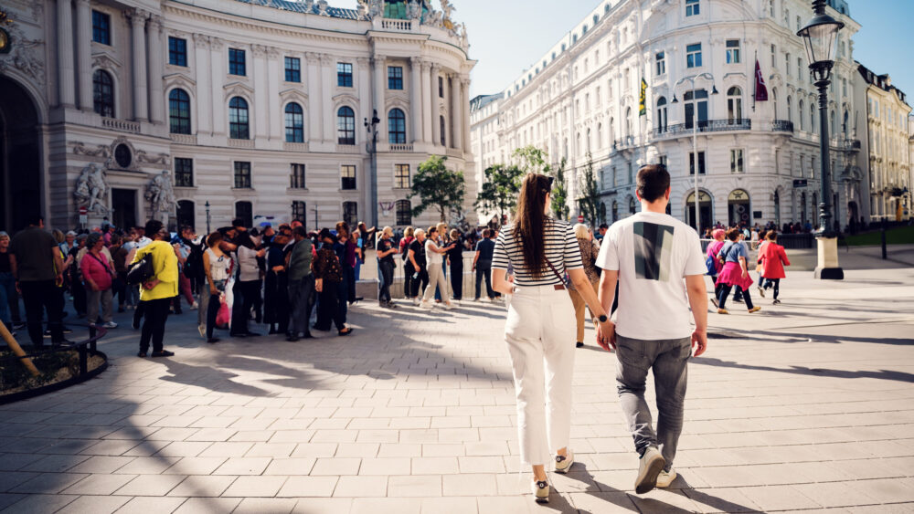 Adriana and Mario holding hands while passing the center of the Michaelerplatz with the imperial architecture surrounding them.