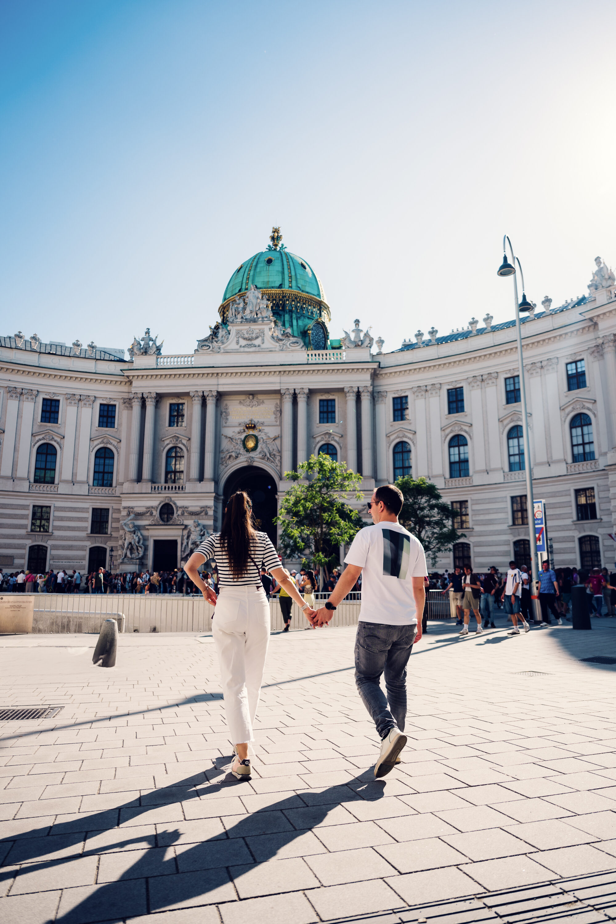 Adriana and Mario strolling through Michaelerplatz square with the Roman excavation site visible in the foreground.