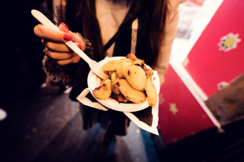 Au marché de Noël, une main aux ongles rouges pose une fourchette sur des rondelles de pommes de terre croustillantes, dans une assiette en carton.