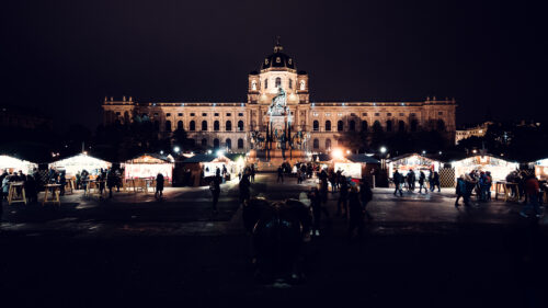 Des chalets et des tables hautes entourent le marché de Noël sur la Maria-Theresien-Platz, et derrière se dressent le monument et le musée illuminés.