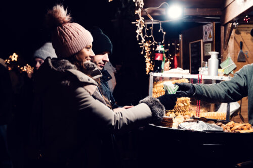 Souriante, Adriana prend un cornet en papier à un stand de pommes de terre en spirale du marché de Noël éclairé d'ampoules, Mario derrière elle.