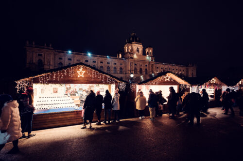 Une lumière chaude se répand des chalets du marché de Noël surmontés d'étoiles sur la Maria-Theresien-Platz, la coupole illuminée du musée en arrière-plan.