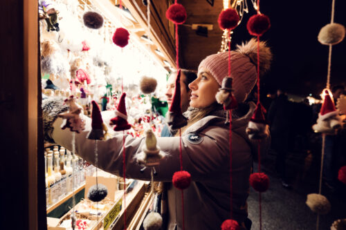 Adriana lève les yeux vers un stand de marché de Noël vivement éclairé, à travers des pompons rouges et des lutins suspendus.