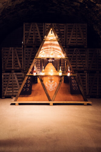 Triangular wooden display installation with Schlumberger bottles and the eagle crest surrounded by stacked crates in the cellar.
