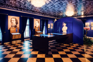 Exhibition room at House of Strauss with portraits, marble bust, and checkered floor.