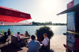 Clienti sulla terrazza di un bar di Copa Beach sotto ombrelloni rossi, lungo il Nuovo Danubio.