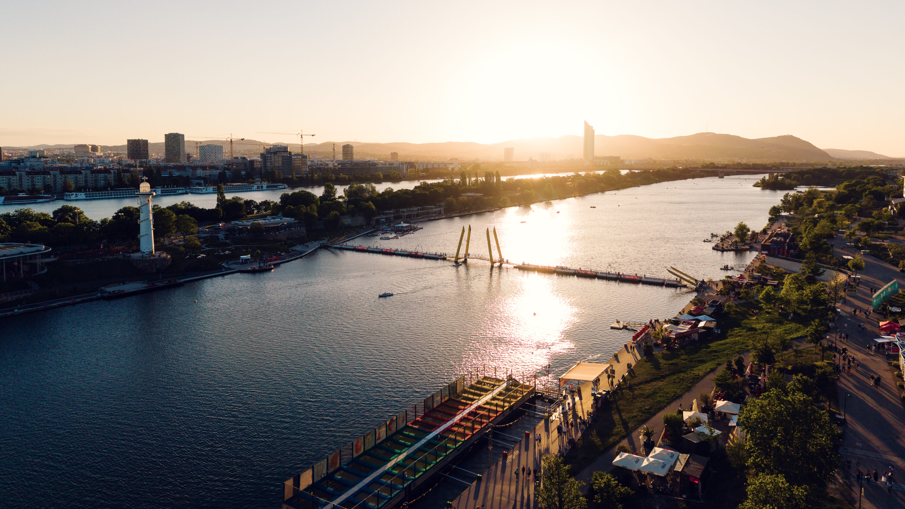 Veduta aerea di Copa Beach nell'ora dorata, con il faro e il Ponte Cagrana sul Nuovo Danubio.