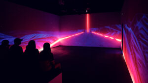 Besucher sitzen im 360-Grad-Projektionsraum vor einer Landschaftsszene mit pinkem Neonlichtstreifen an den Wänden.