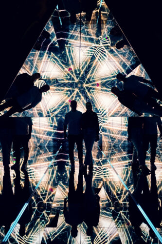Silhouette von Adriana und Mario im verspiegelten Pyramidenraum, umgeben von goldenen geometrischen Lichtmustern.