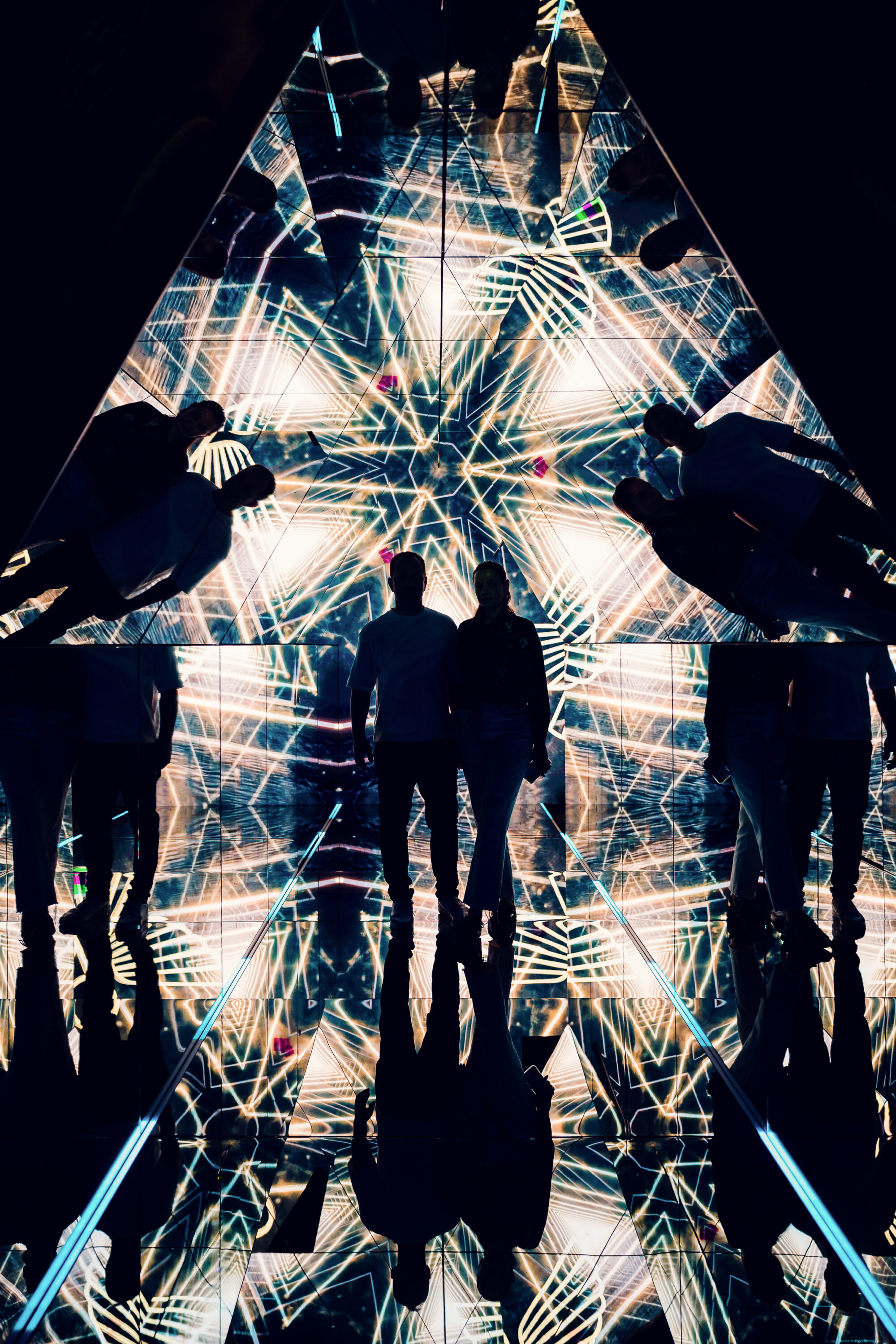 Silhouette von Adriana und Mario im verspiegelten Pyramidenraum, umgeben von goldenen geometrischen Lichtmustern.