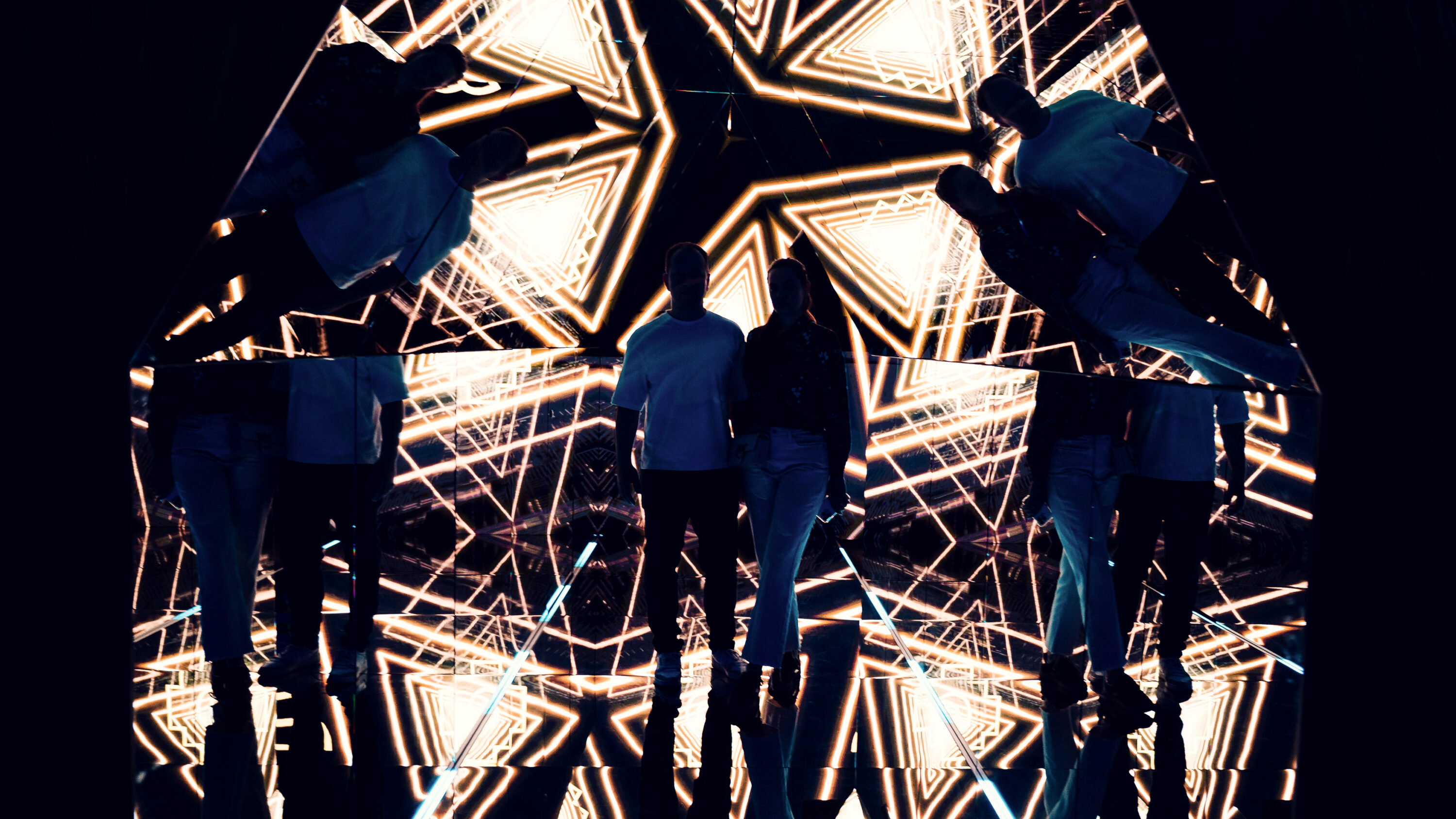 Two visitors standing in the pyramid room with geometric golden projections reflected around them.