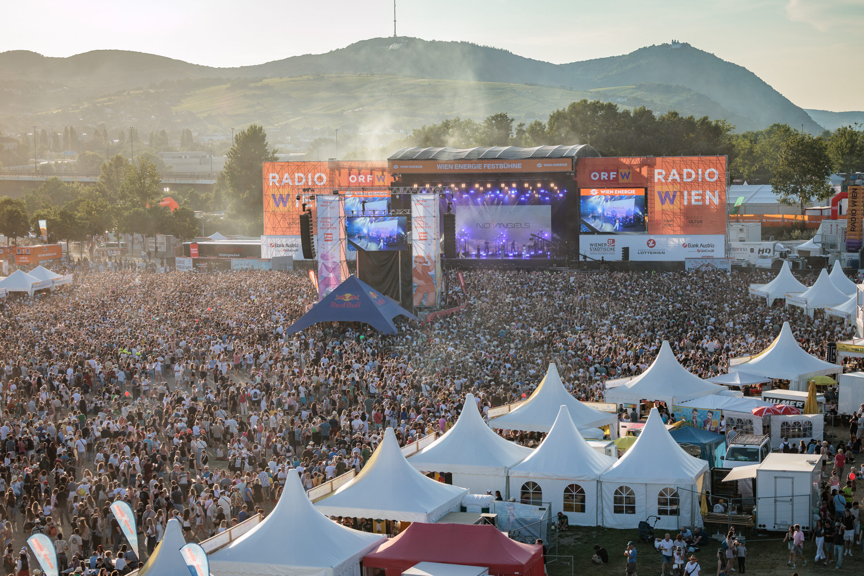 Daytime crowd in front of the Wien Energie main stage at Donauinselfest with festival tents and the Vienna woods behind